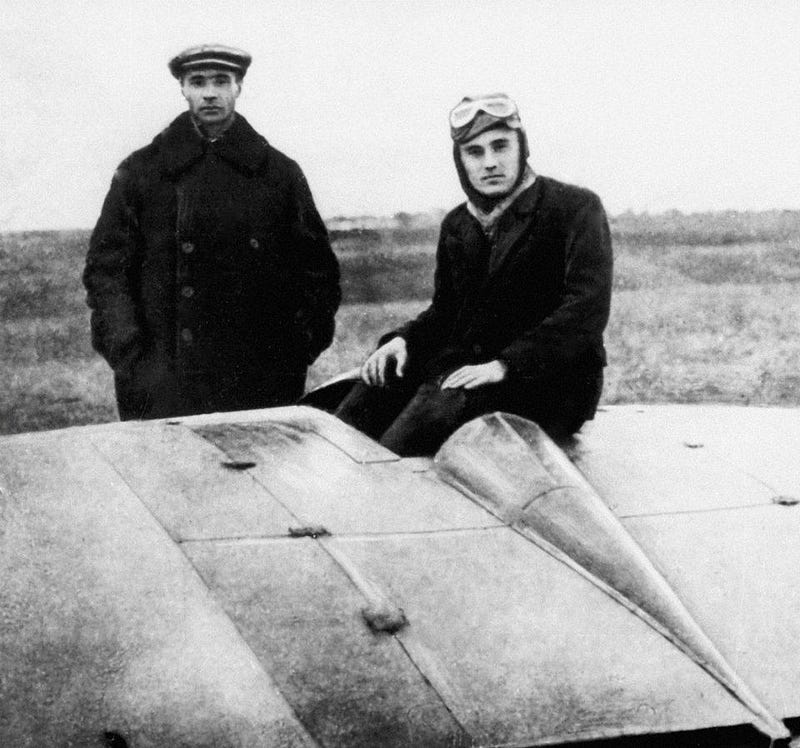 Two men pose on and beside an early 20th-century aircraft, reminiscent of pioneering days that eventually led to moon endeavors. One stands as the other sits on the plane, wearing goggles, embodying the spirit of exploration in the USA and beyond. The background reveals an open field.