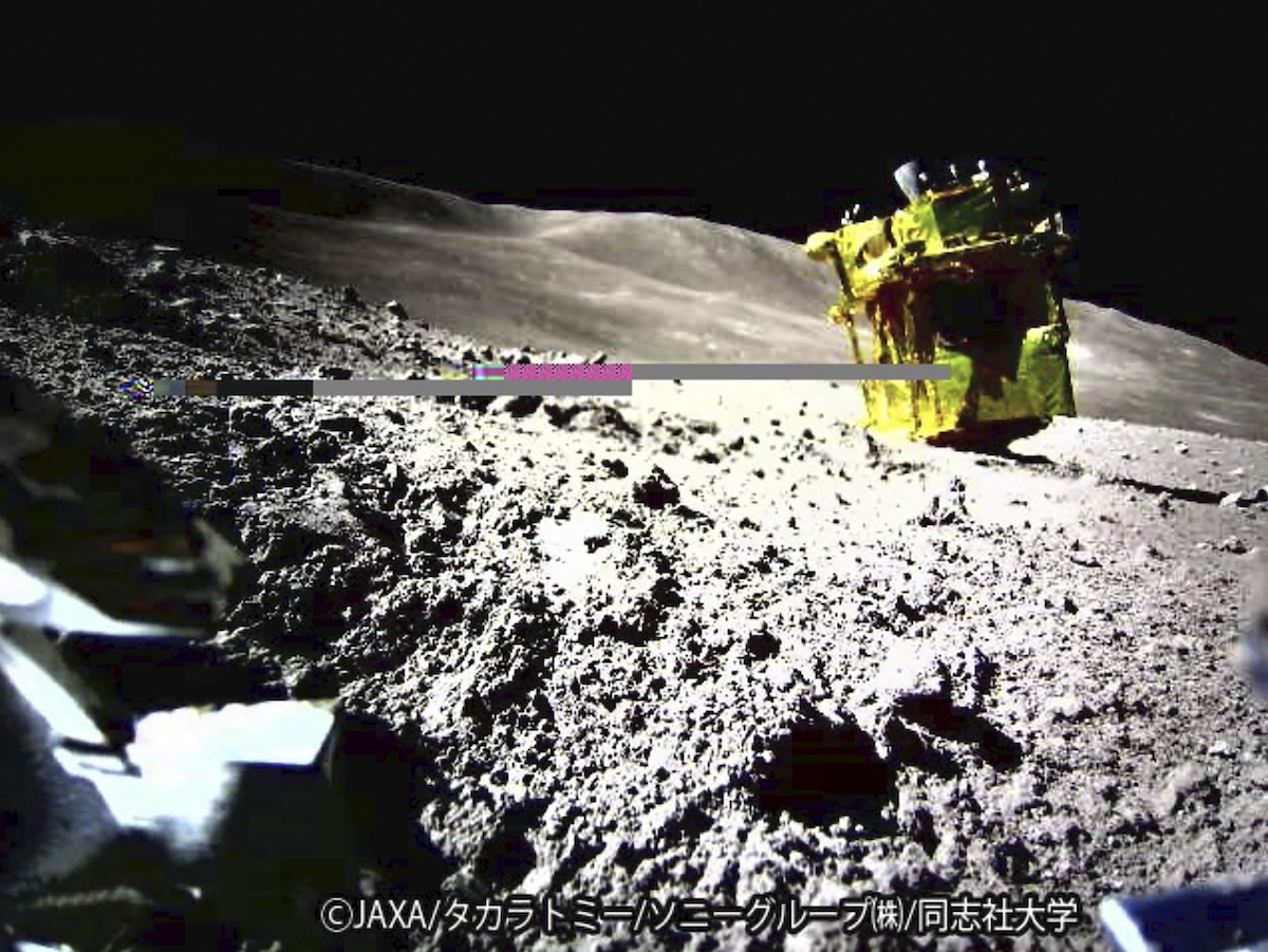 A yellow spacecraft, possibly the Hayabusa2 lander, is perched on a rocky surface of an asteroid, with rough terrain visible under a clear sky. Visible text includes &copy;JAXA and Japanese characters.