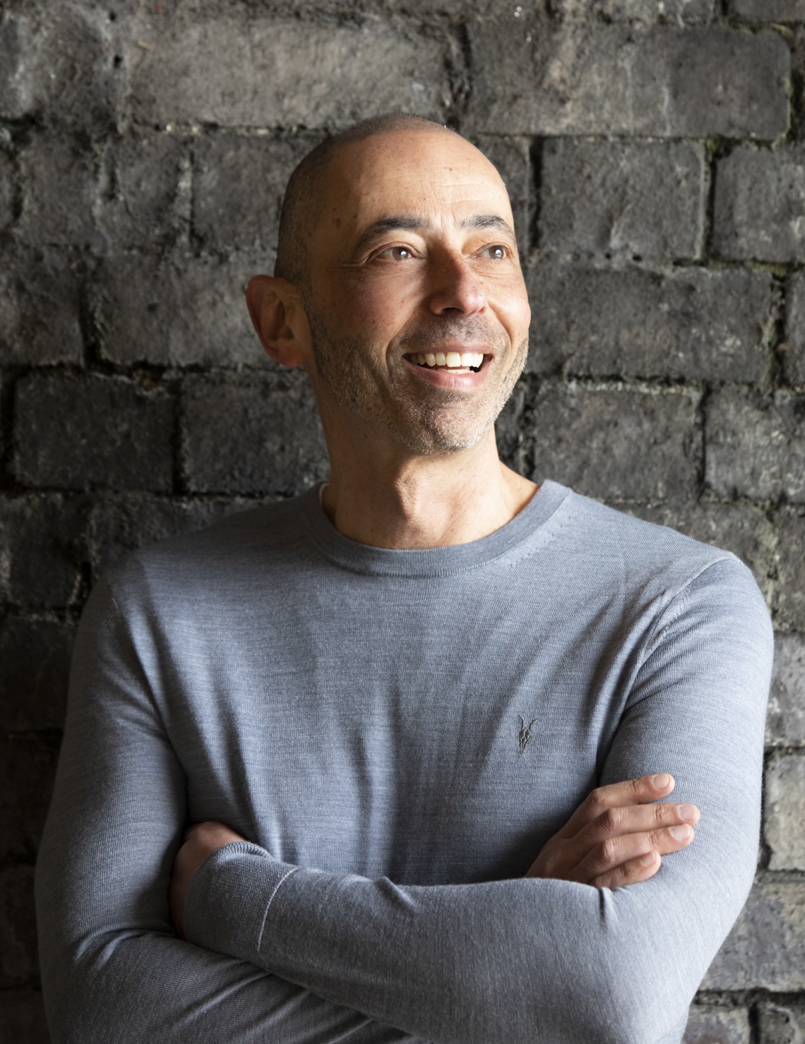 A person in a gray sweater stands against a brick wall, smiling with arms crossed.
