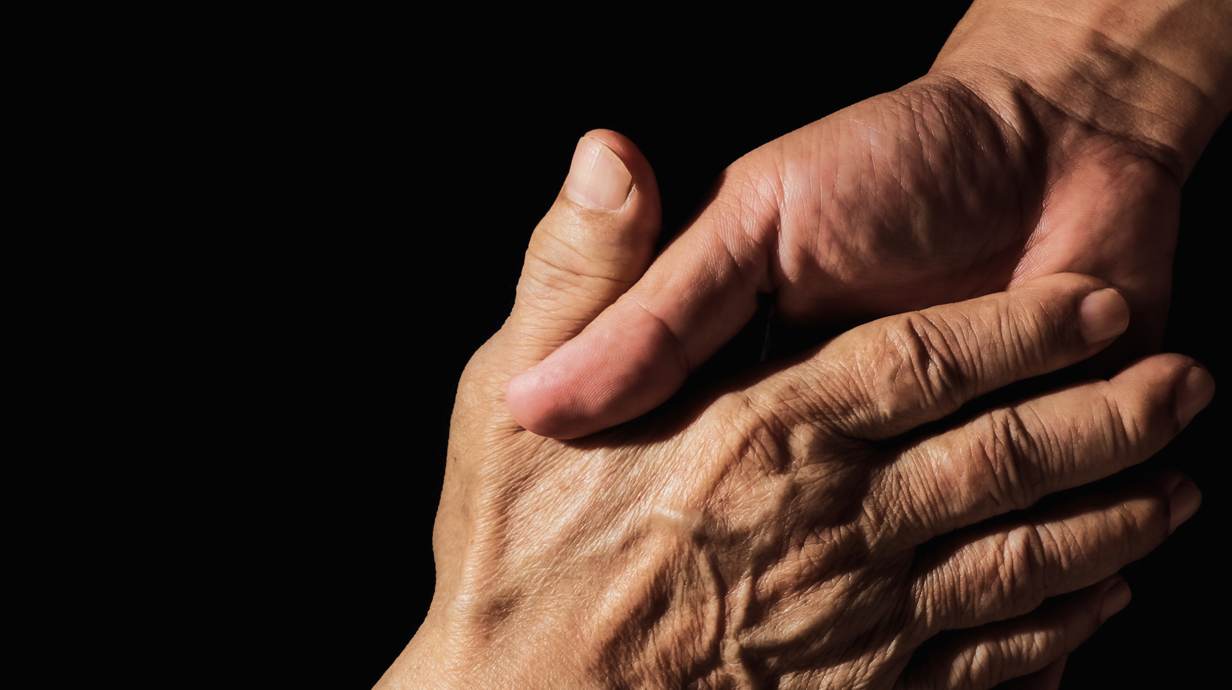 A hand clasps an older, wrinkled hand against a black background, symbolizing care and support.