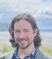 Man with long hair and beard smiling outdoors, wearing a leather jacket and plaid shirt, with a cloudy sky and water in the background.