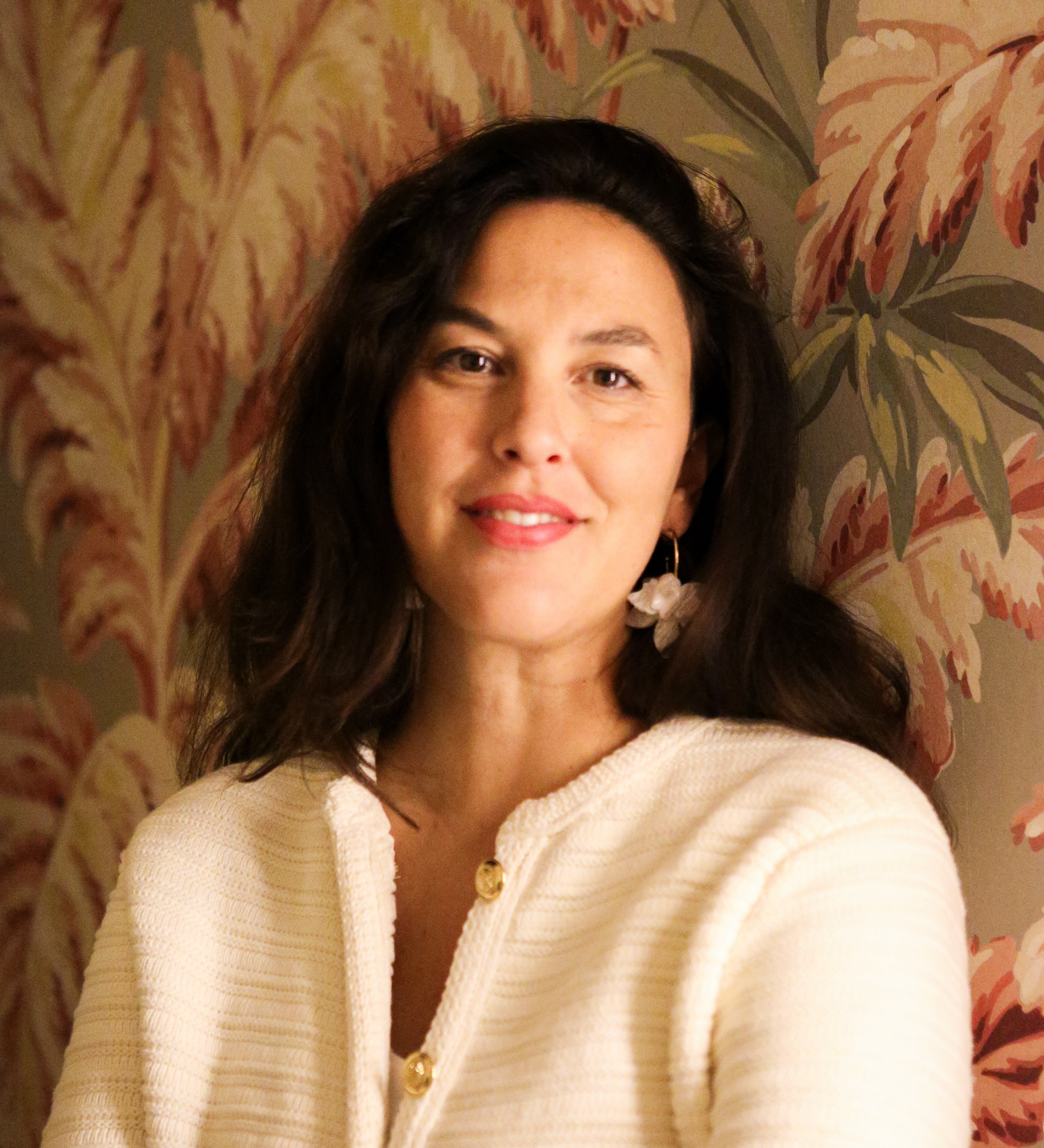 A person with long dark hair smiles, wearing a light-colored top and earrings, standing against a patterned wallpaper with leaf designs.