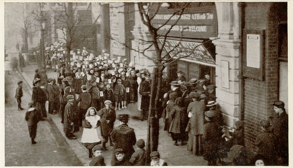 A large group of people, including children, line up outside a historic building on a city street.