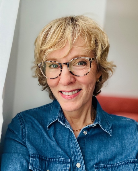A woman with short blonde hair and glasses smiles while wearing a denim shirt.