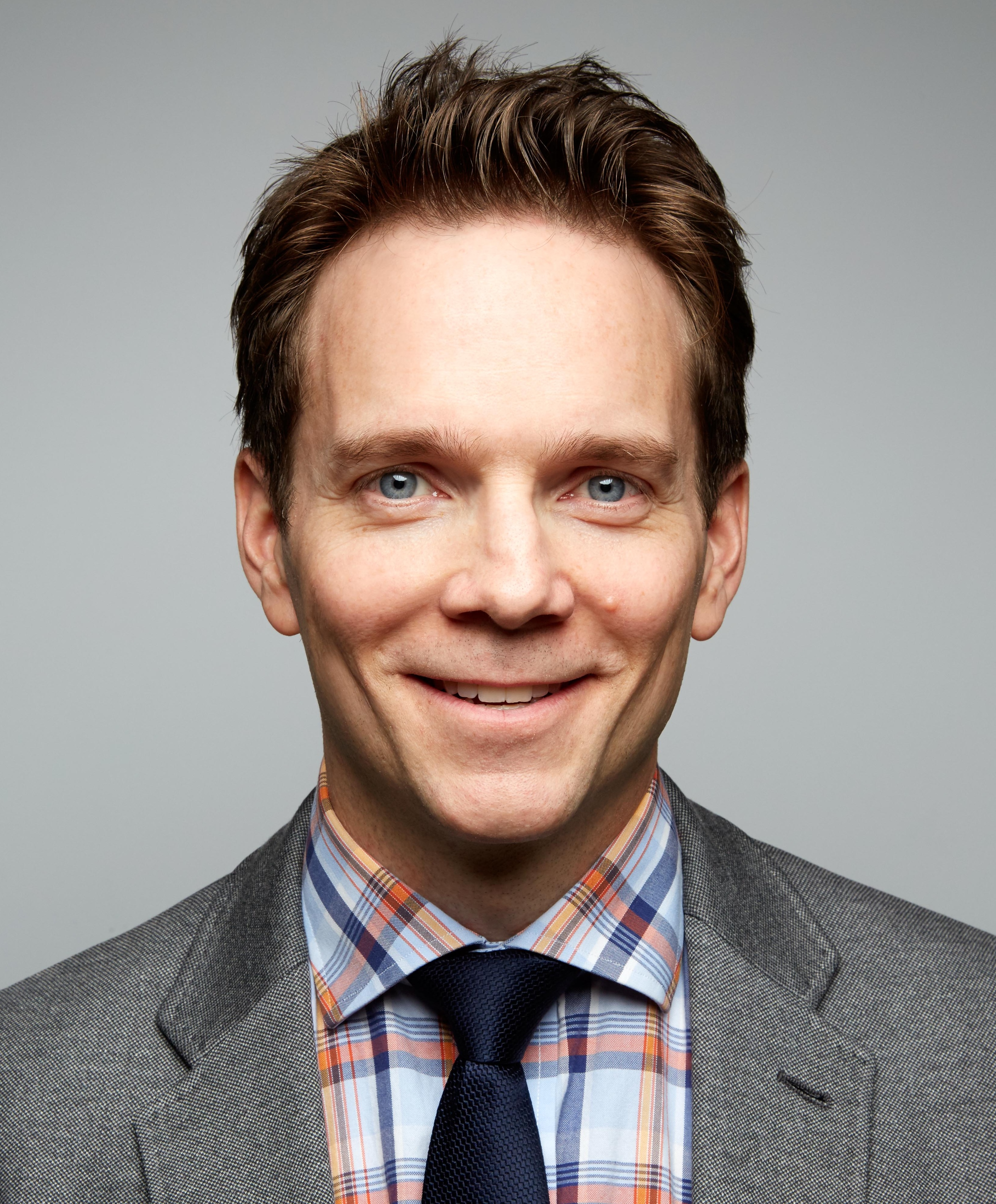 A man in a gray suit and checkered shirt with a dark tie smiles at the camera against a plain background.