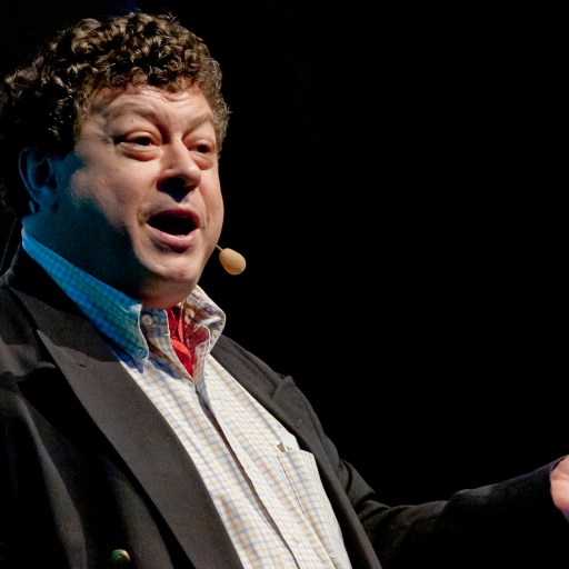 A man with curly hair wearing a suit and headset microphone gestures while speaking on stage.