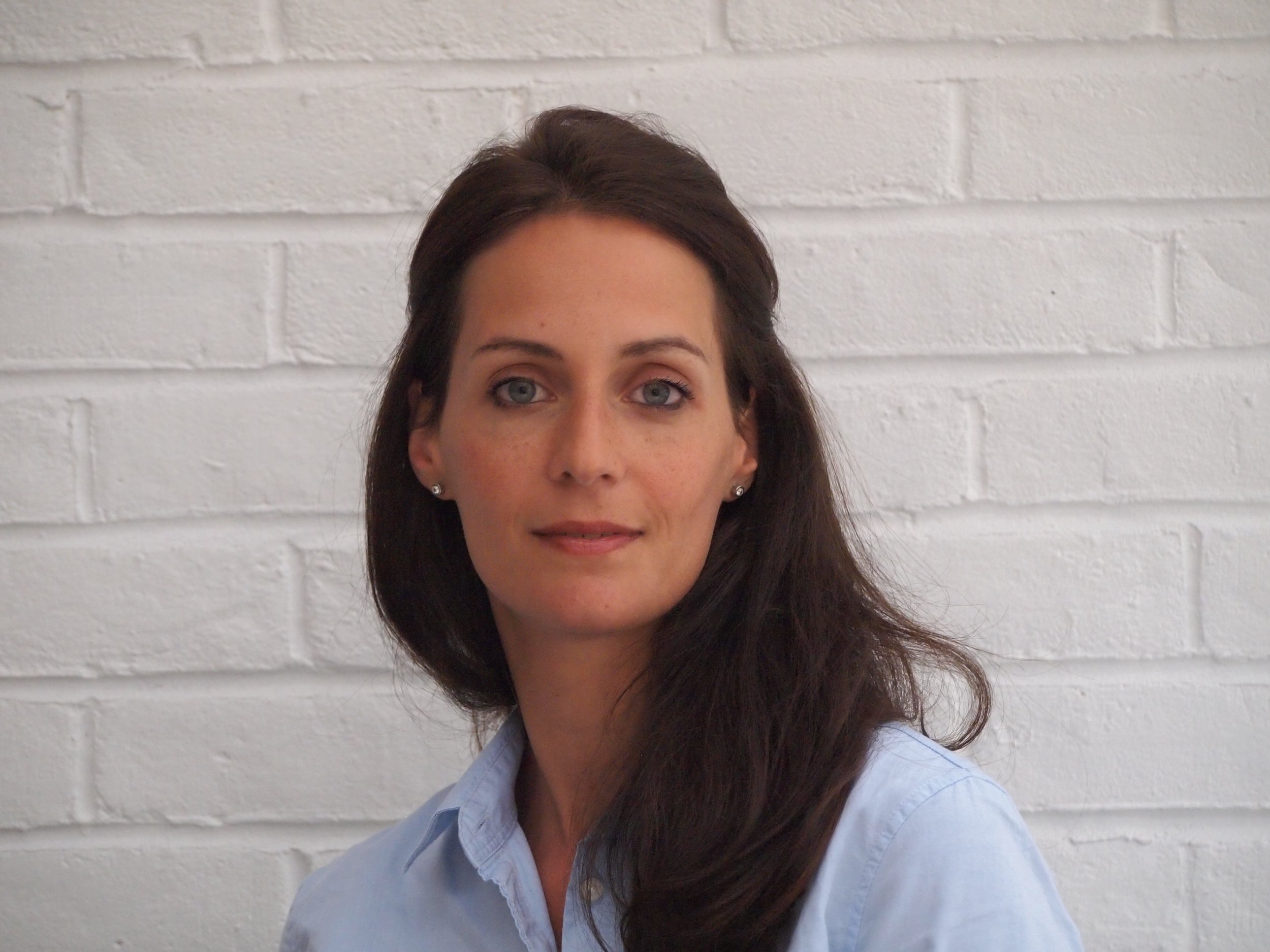A woman with long dark hair and a blue shirt stands against a white brick wall.