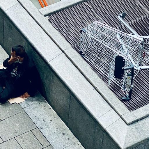 A person sits on a sidewalk near an escalator entrance, next to an overturned shopping cart on a metal grate.
