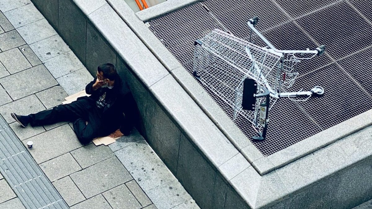 A person sits on a sidewalk near an escalator entrance, next to an overturned shopping cart on a metal grate.