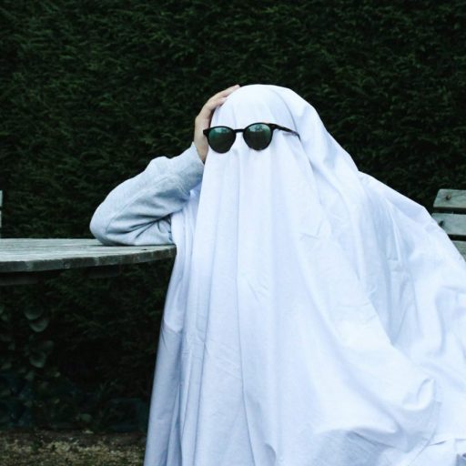 Person disguised as a ghost with sunglasses, sitting at an outdoor table.