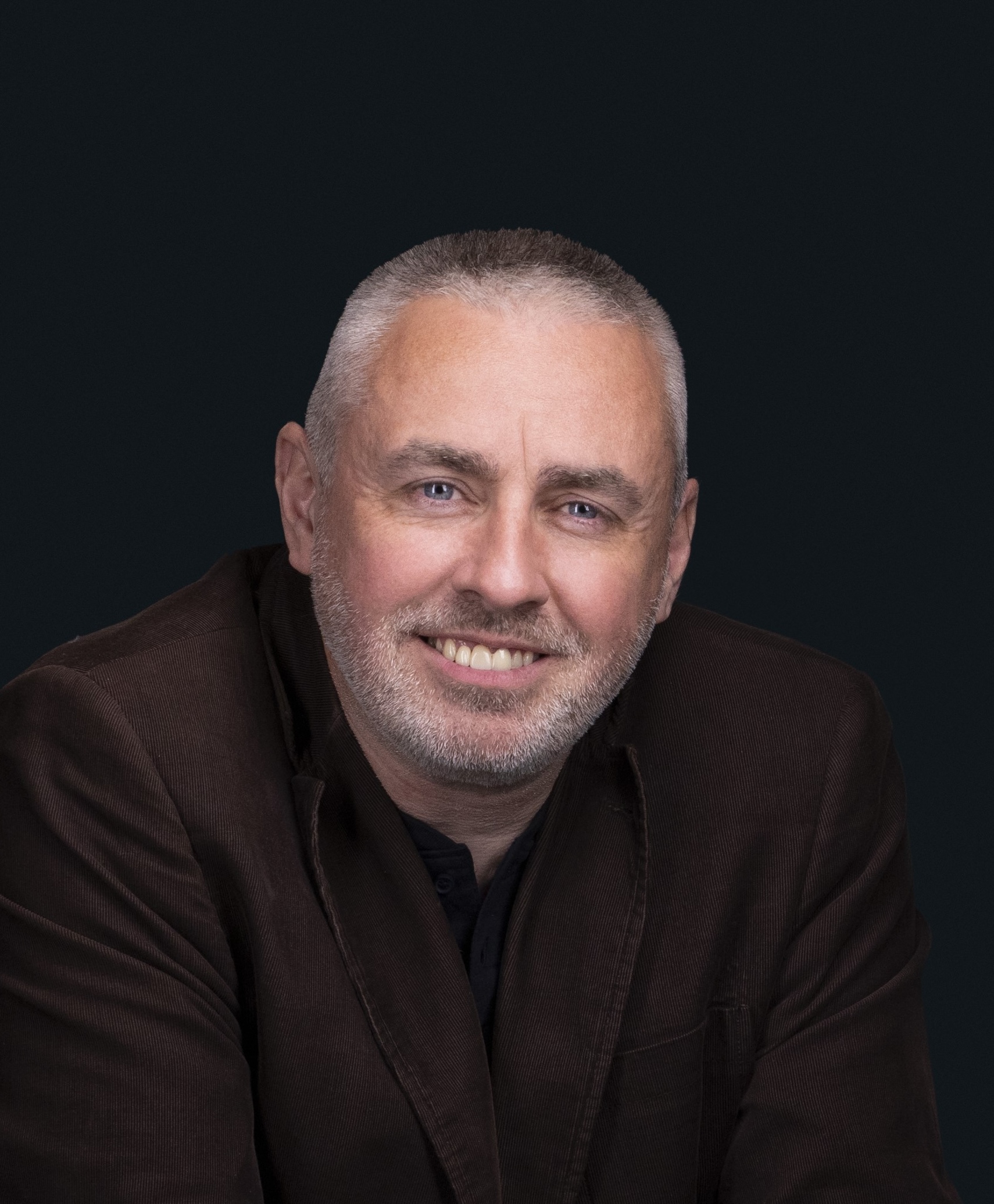 A man with short gray hair and a beard is smiling, wearing a dark blazer against a solid dark background.