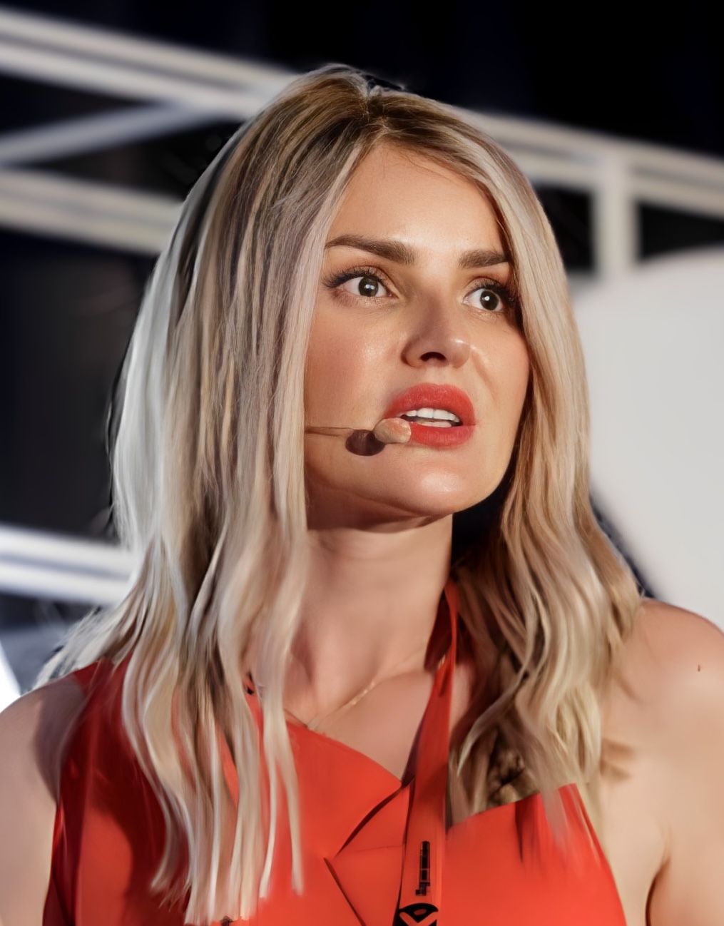 A woman with long blonde hair is speaking into a headset microphone, wearing a red top.