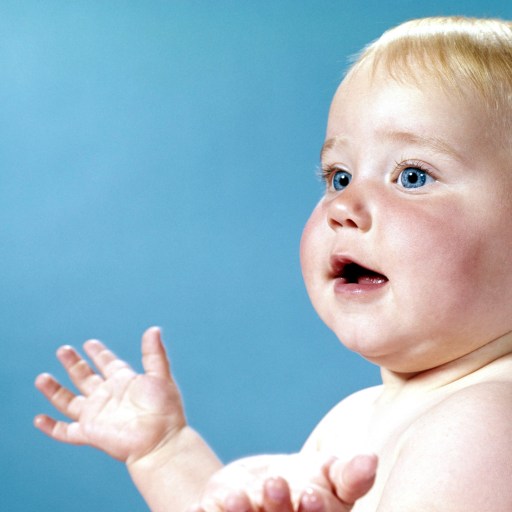 A baby with blonde hair and blue eyes looks upward with a curious expression against a blue background.