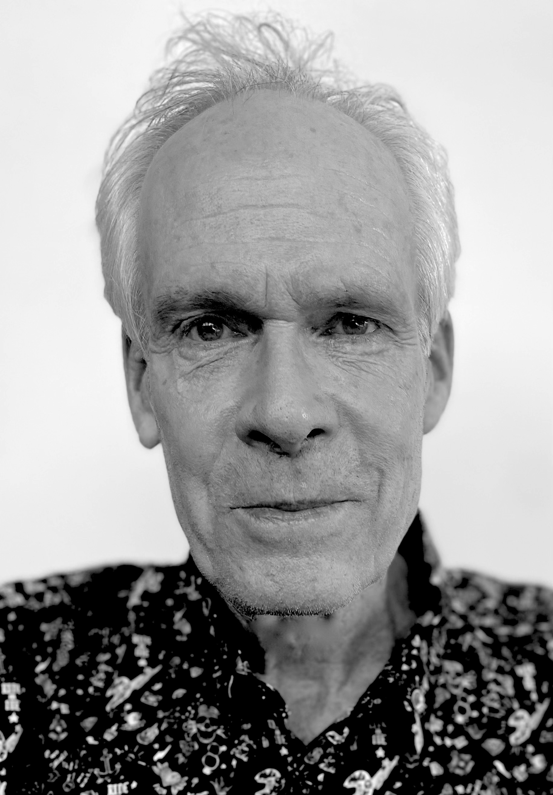 Black and white portrait of an older man with a neutral expression, wearing a patterned shirt, against a plain background.