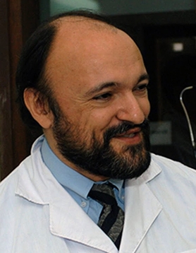 A person with a beard and short hair, wearing a white coat over a blue shirt and tie, smiles while looking to the side.