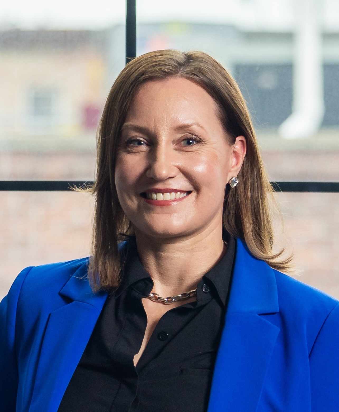 A person with shoulder-length brown hair is smiling, wearing a blue blazer over a black shirt, posed in front of a window.
