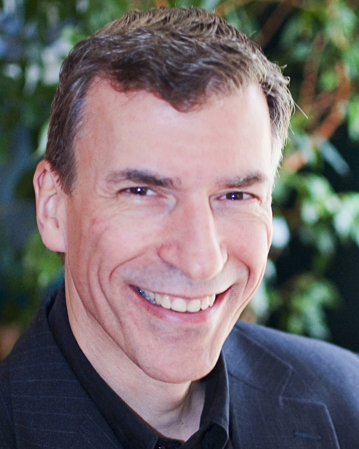 A smiling man in a suit stands in front of a blurred green background.