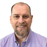 A man with a beard is smiling, wearing a light purple, checkered shirt. The background is plain white.