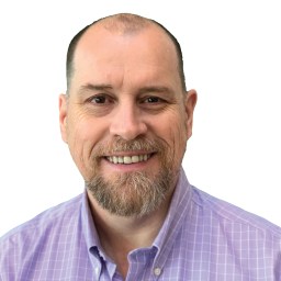 A man with a beard is smiling, wearing a light purple, checkered shirt. The background is plain white.