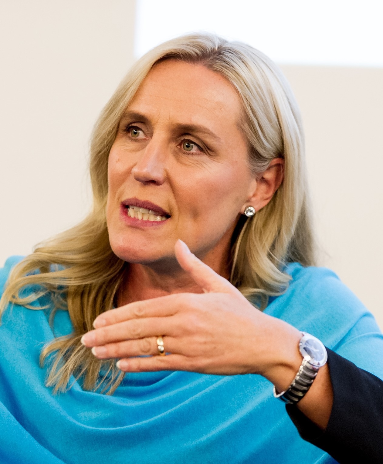 A woman with long blonde hair gestures while speaking. She is wearing a blue top, earrings, and a watch.