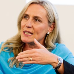 A woman with long blonde hair gestures while speaking. She is wearing a blue top, earrings, and a watch.