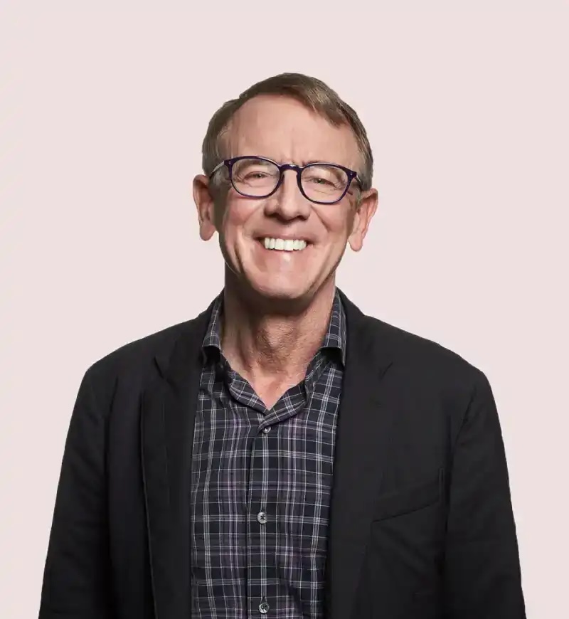 A smiling person with glasses and short hair, wearing a checkered shirt and dark blazer, stands against a plain background.