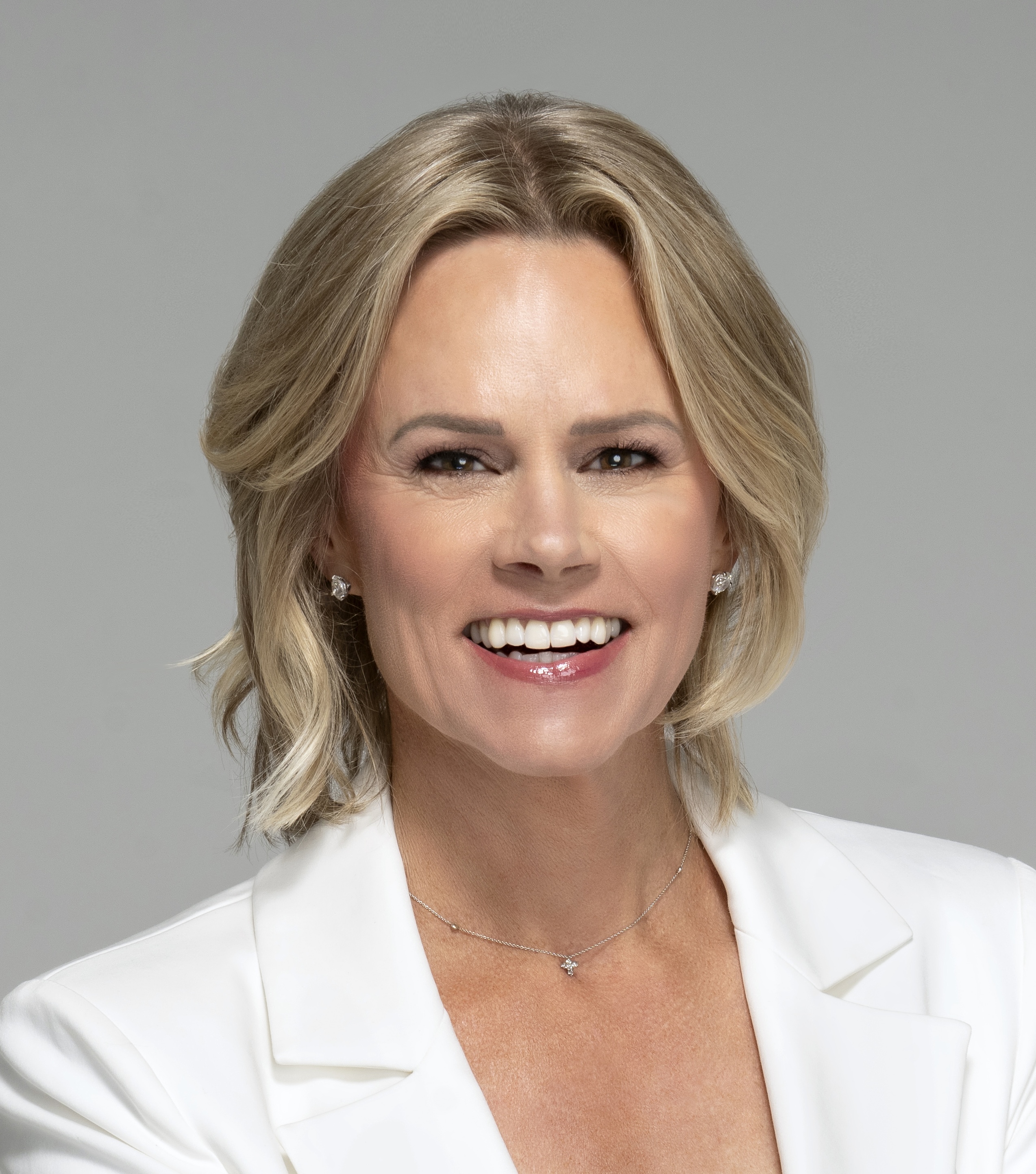 A person with shoulder-length blonde hair, smiling, wearing a white blazer and necklace, against a neutral background.