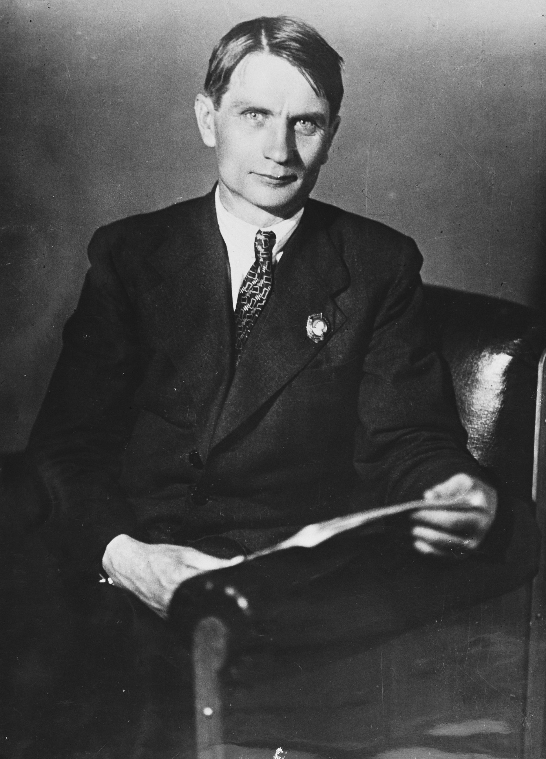 In a black and white portrait, a man in a suit sits on a chair, thoughtfully holding a paper. His expression suggests contemplation, seemingly reflecting on the impact of government decisions or advancements in science.