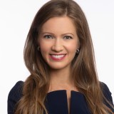 A woman with long brown hair smiles, wearing a dark blue outfit.
