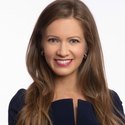 A woman with long brown hair smiles, wearing a dark blue outfit.