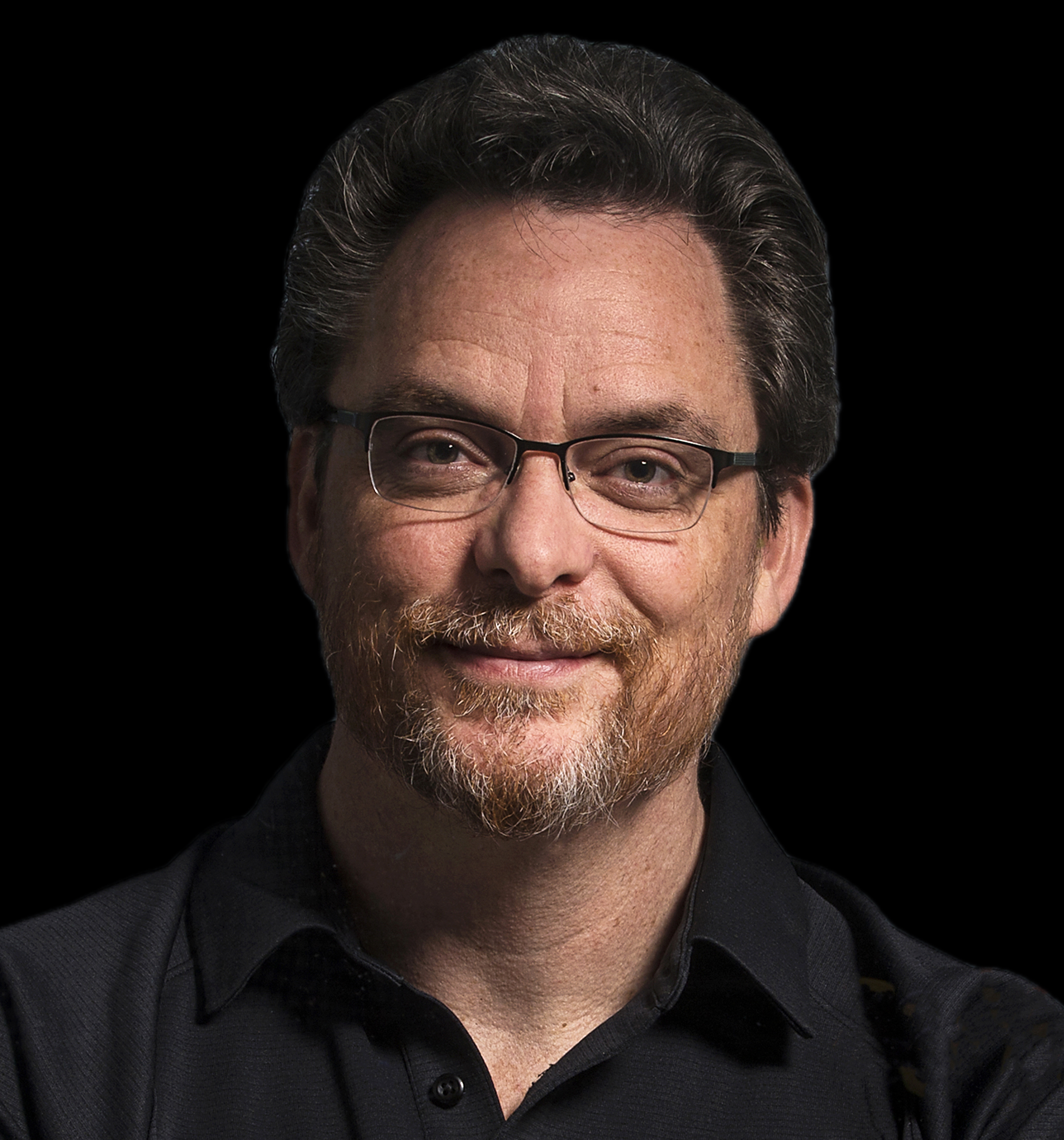 A person with glasses and a beard smiles against a black background, wearing a dark shirt.