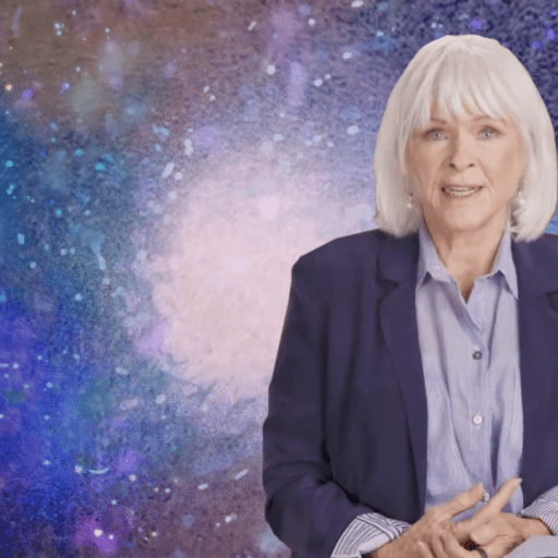 A person with white hair sits in front of a colorful, abstract background. They are wearing a dark blazer over a striped shirt.