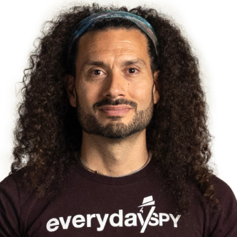 Man with curly hair, wearing a headband and "everyday spy" t-shirt, standing against a white background.