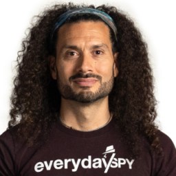 Man with curly hair, wearing a headband and "everyday spy" t-shirt, standing against a white background.