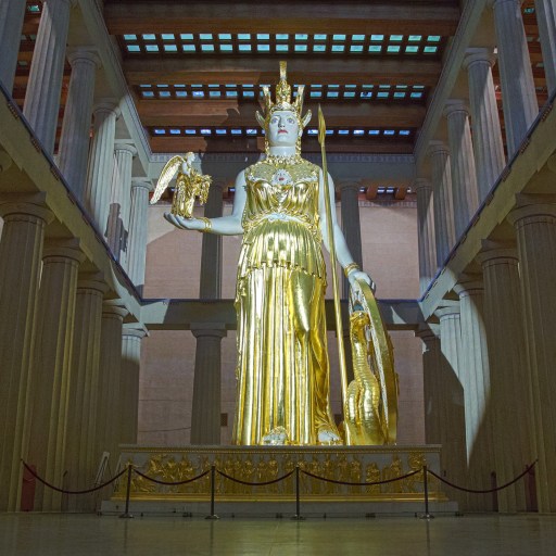The golden statue of Athena stands prominently in a replica of the Parthenon interior, steeped in mythology. She holds a shield and a small figure, while the spacious setting boasts large columns and high ceilings.