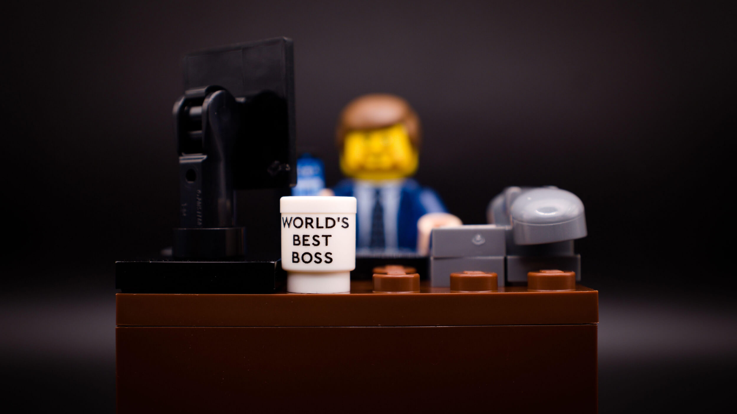 LEGO minifigure dressed in a suit sits at a desk with a computer monitor, phone, and a mug labeled "World's Best Boss.
