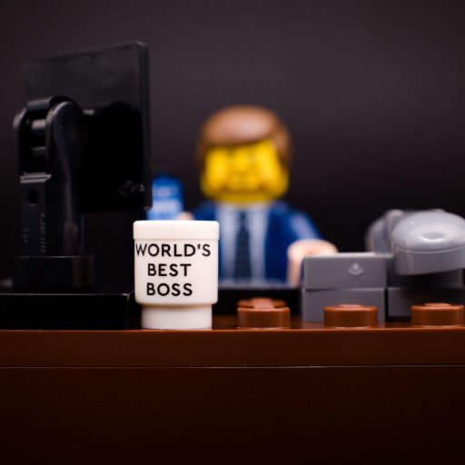 LEGO minifigure dressed in a suit sits at a desk with a computer monitor, phone, and a mug labeled "World's Best Boss.