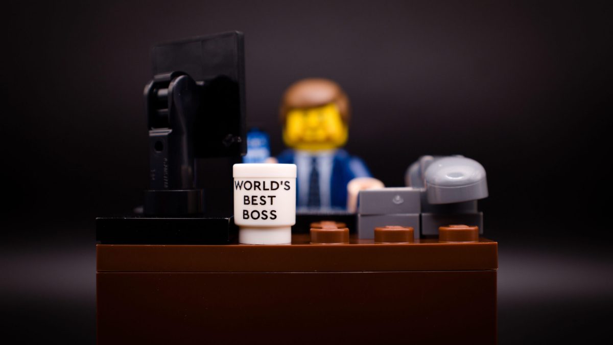 LEGO minifigure dressed in a suit sits at a desk with a computer monitor, phone, and a mug labeled "World's Best Boss.