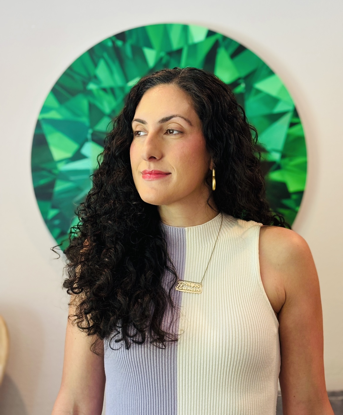 A woman with long curly hair stands in front of a green geometric background, wearing a sleeveless two-tone top and a necklace.