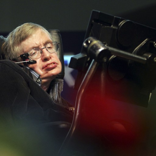 A person seated in a wheelchair uses a communication device with a Stephen Hawking-like voice, blurred flowers gently framing the scene in the foreground.