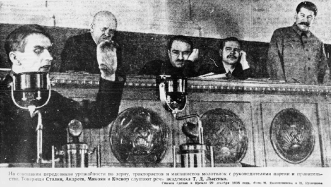 Five men in formal attire, embodying both the spirit of government and scientific inquiry, observe a speech from a raised platform; microphones are carefully positioned on the railing in front of them.