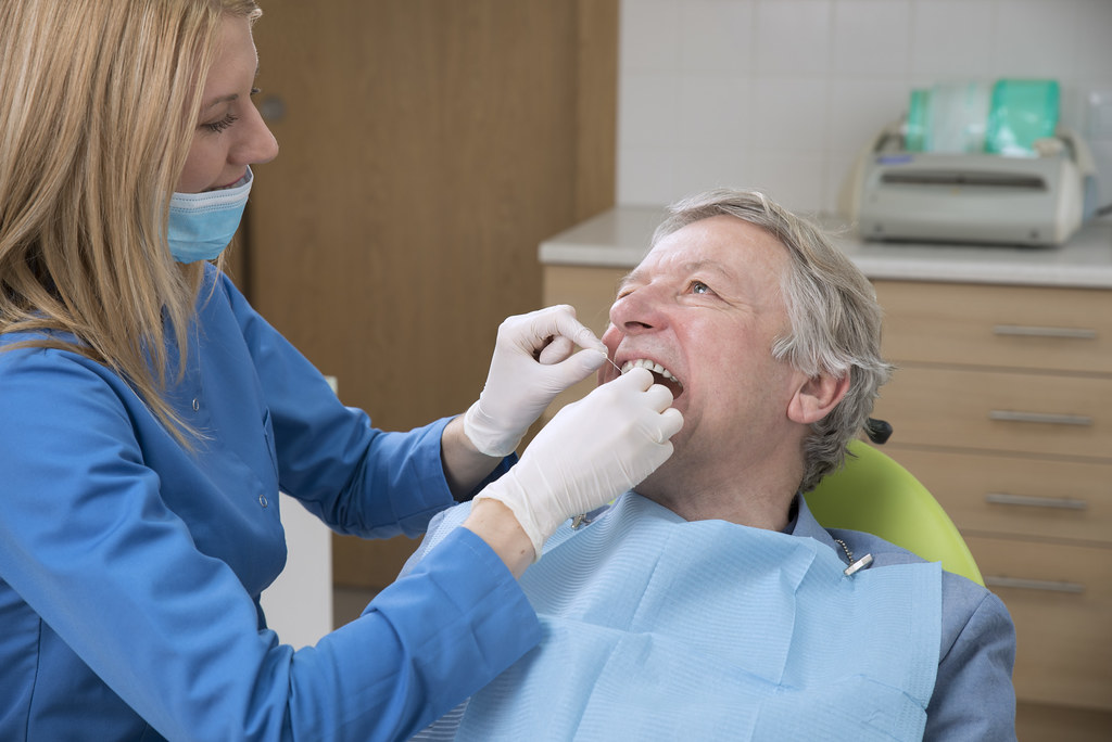 A dental professional examines an older man's teeth in a dental clinic. They both wear protective gear.