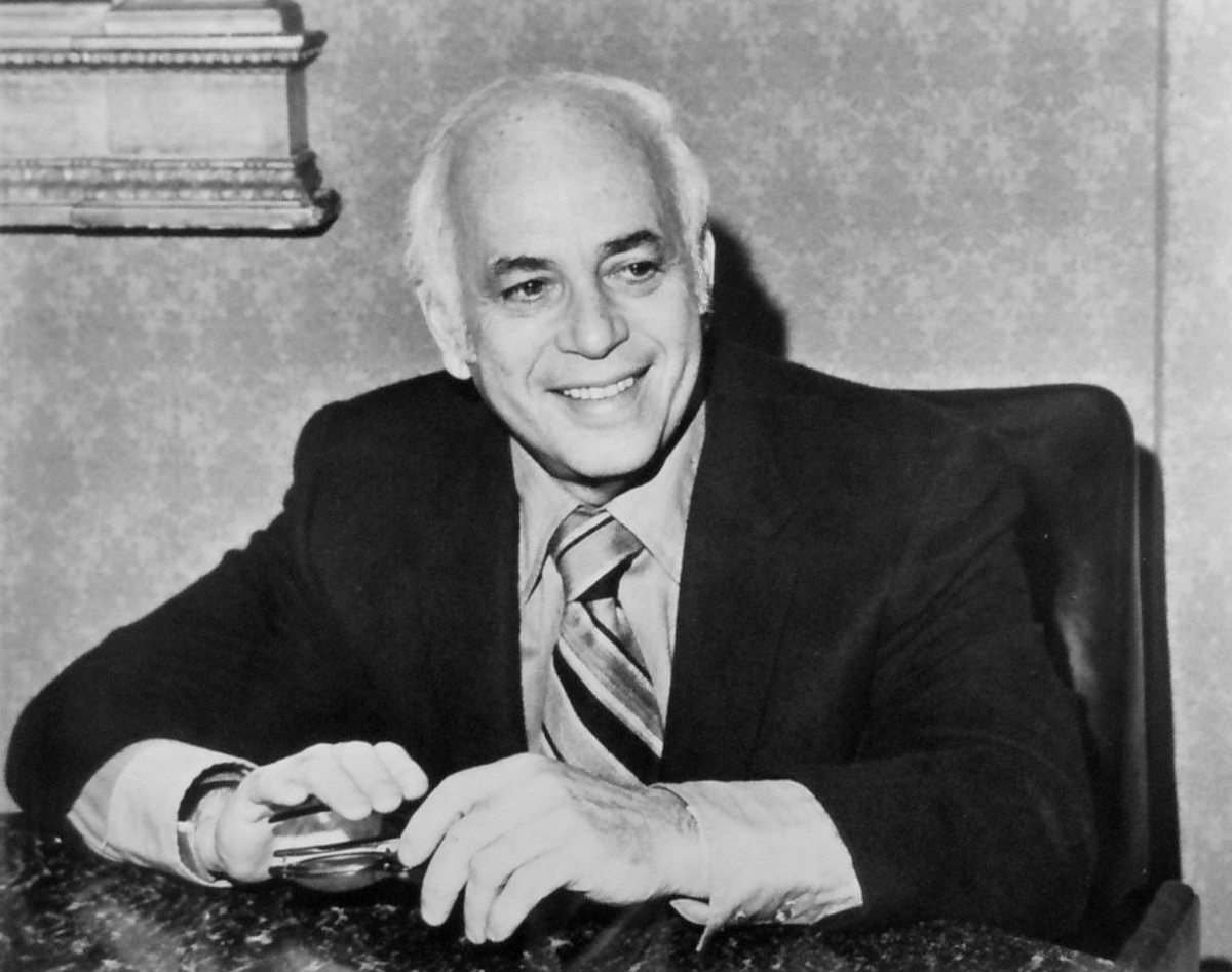 A man in a suit and tie sits at a desk, smiling, with hands resting on the table.