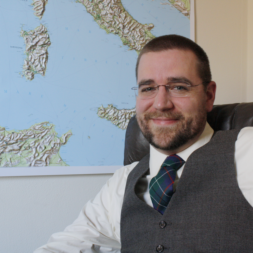 A man in glasses, wearing a vest and tie, sits before a map of Italy on the wall, lost in thought as if planning a Martian Revolution.