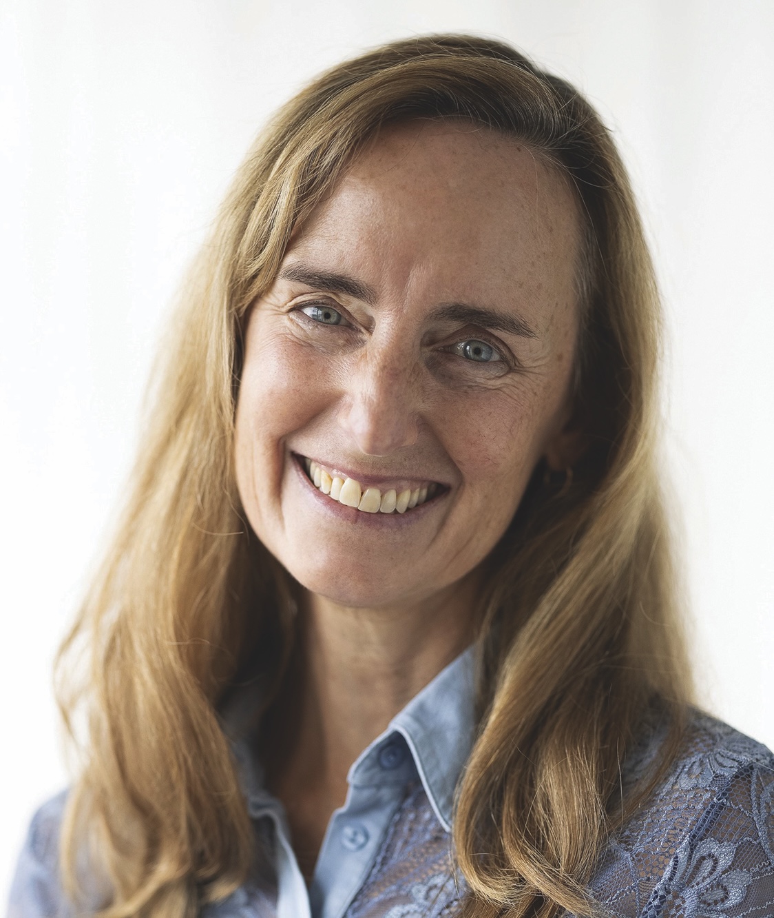 A woman with long hair smiles while wearing a blue blouse against a neutral background.