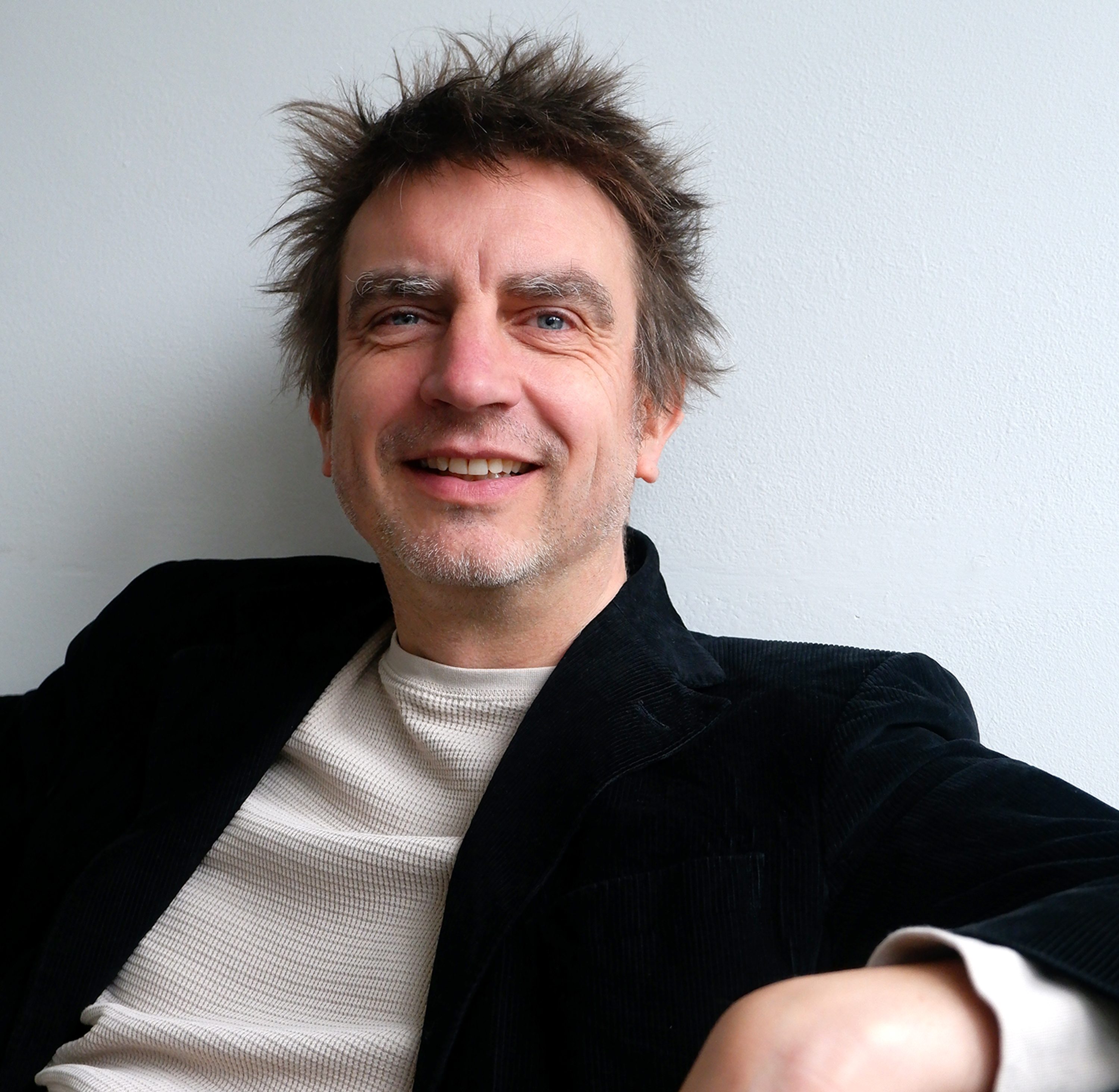 A man with tousled hair, wearing a black jacket and light sweater, sits against a white wall, smiling at the camera.