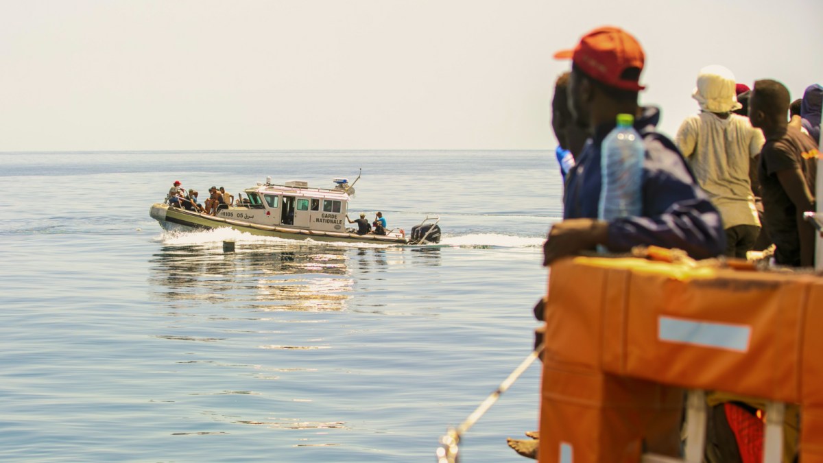 A group of people on a boat watch as a patrol vessel approaches on calm, open water under a clear sky, reflecting ongoing migration statistics in the region.