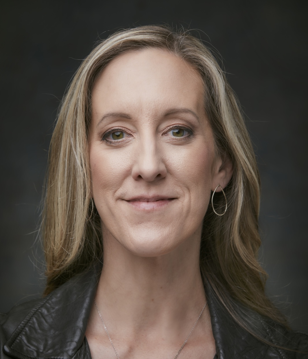 A person with long blonde hair and a leather jacket smiles slightly against a dark background.