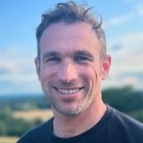 A man with short hair and a beard smiles at the camera, standing outdoors with a blurred landscape and blue sky in the background.