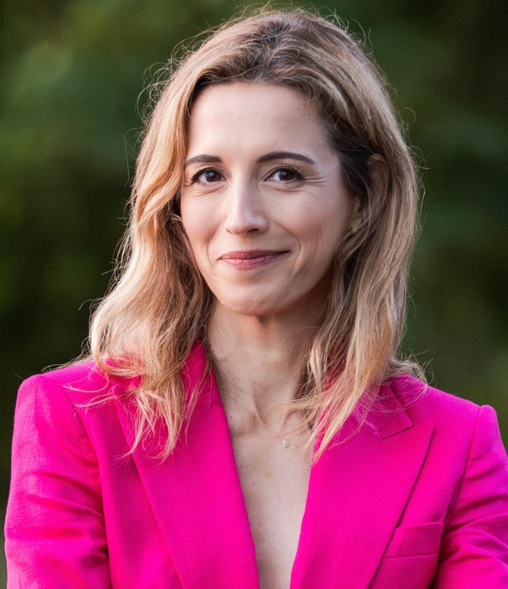 A woman with shoulder-length light brown hair wearing a bright pink blazer smiles at the camera. The background is blurred greenery.